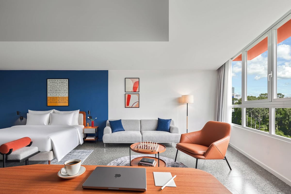 A modern hotel room with a bed, a sofa, a chair, a desk with a laptop, and a view of the outdoors.