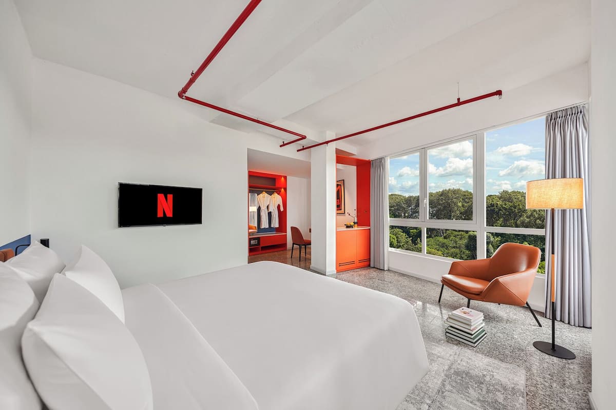 A modern hotel room with a large bed, a TV mounted on the wall, a wardrobe, a seating area with a chair, and a view of the outdoors through large windows.