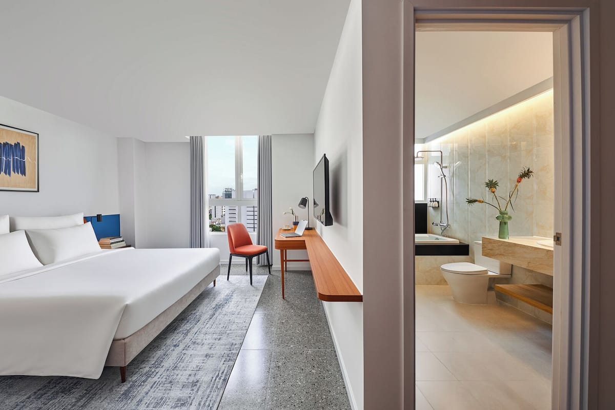 A modern hotel room with a large bed, a desk, a chair, and a bathroom with a shower and toilet.
