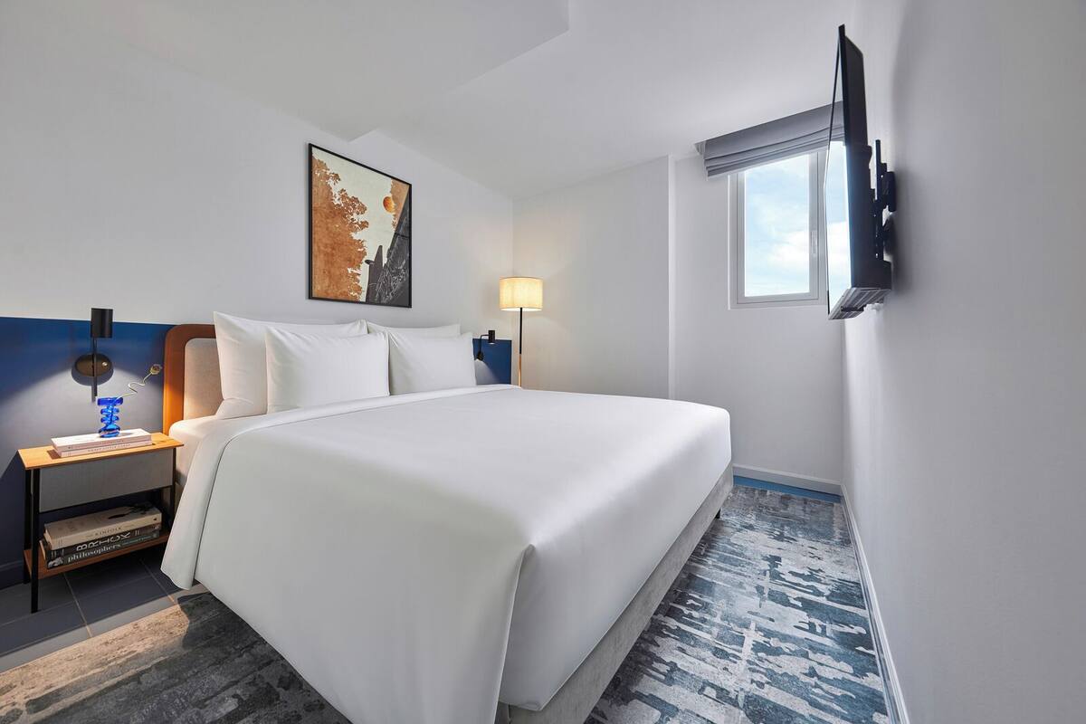 A hotel room with a large bed, a nightstand with a lamp, a painting on the wall, and a TV mounted on the wall.