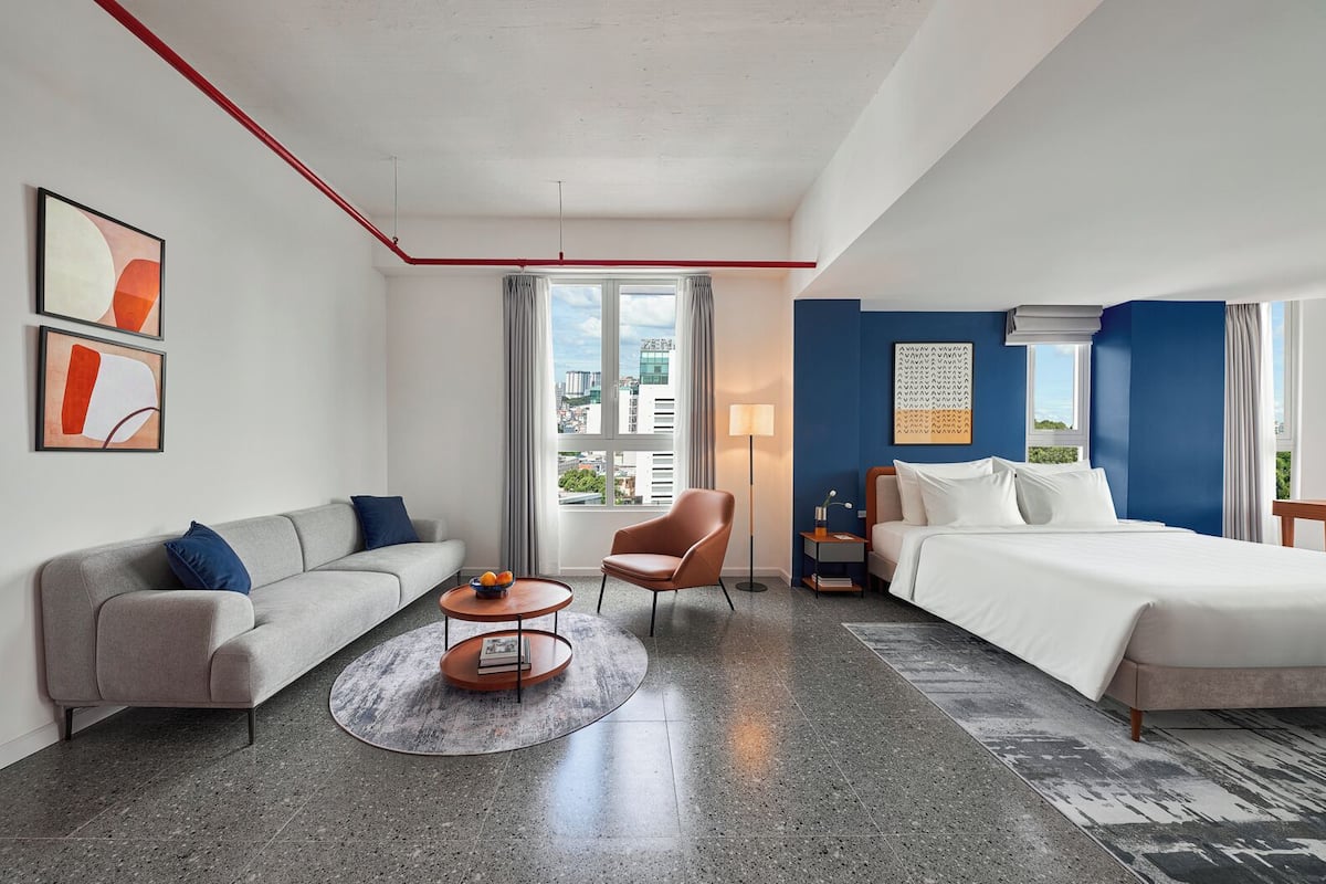 A hotel room with a bed, a sofa, a chair, a small table, and a lamp. There is a window with a view of buildings.