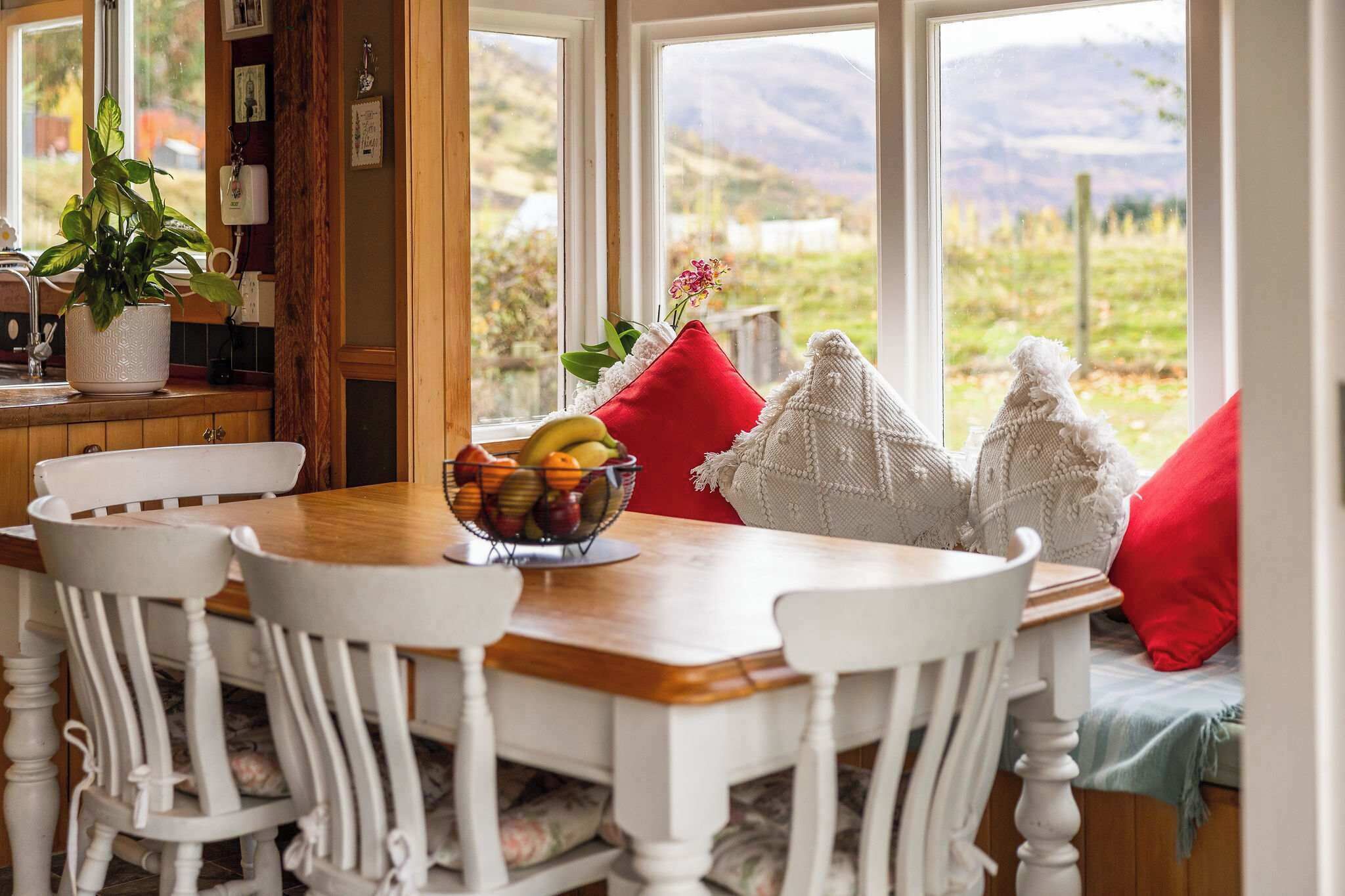 A dining area with a wooden table, white chairs, a bowl of fruit, and a view of the outdoors through large windows.