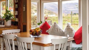 Un comedor con una mesa de madera, sillas blancas, un cuenco de fruta y vistas al exterior a través de amplias ventanas.