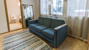 Ein Zimmer mit einem blauen Sofa, einem Streifenmuster-Teppich, einer Holzbalkendecke und einem Fenster mit Blick auf Bäume.