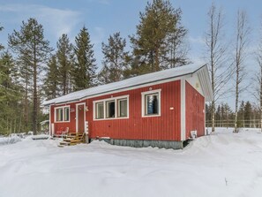 Une cabane rouge, dont le toit est recouvert de neige, entourée d’arbres et s’inscrivant dans un paysage hivernal.