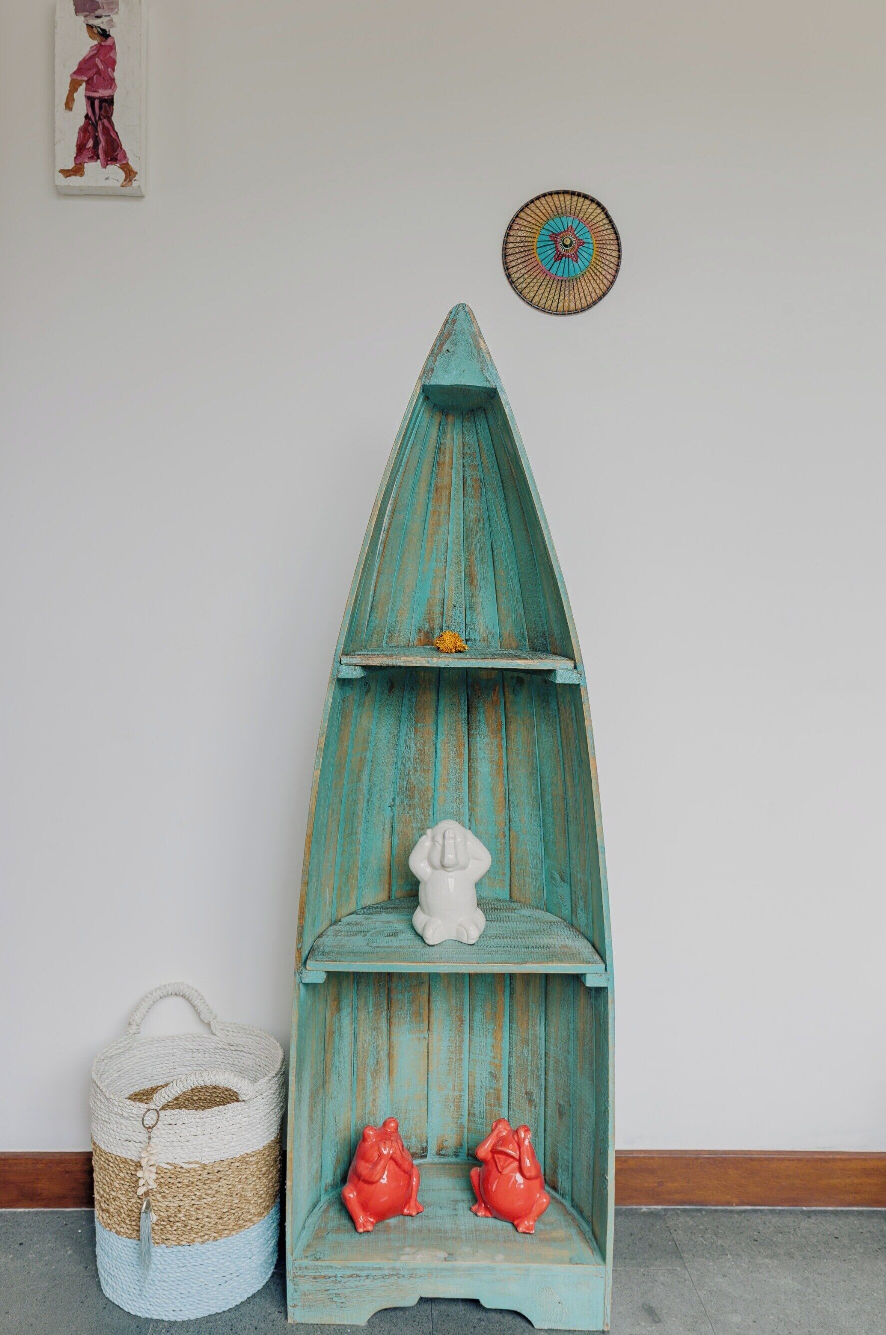 Une étagère en bois de couleur turquoise, ornée de deux grenouilles rouges, d’une figurine blanche et d’un panier tressé, le tout placé contre un mur uni.