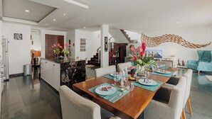 A spacious dining area with a wooden table set for four, surrounded by comfortable chairs. A kitchen area is visible to the left, and a staircase leads to an upper level.
