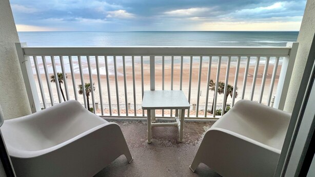 Ein Balkon mit zwei weißen Stühlen und einem kleinen Tisch mit Blick auf einen Strand, der mit Palmen und einem bewölkten Himmel gesäumt ist.
