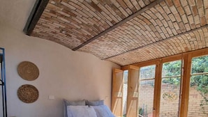 Ein Schlafzimmer mit Ziegeldecke, einem Bett mit weißer Bettwäsche, Holzboden und Blick durch ein Fenster.