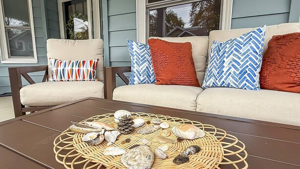 A cozy outdoor seating area with a wicker table displaying seashells and a pine cone, a beige sofa with patterned pillows, and a wooden chair.