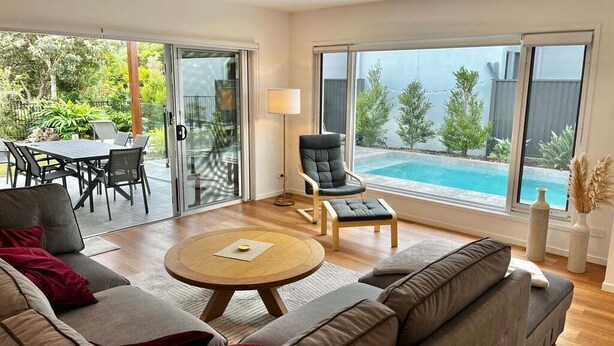 Una sala de estar moderna con un sofá, una mesa de centro redonda y una silla. Hay una mesa de comedor con sillas afuera de una puerta de vidrio. Un ventanal ofrece vistas a una piscina.