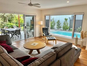 Un salon moderne avec un canapé, une table basse ronde et un fauteuil. Dehors, derrière une porte vitrée, se trouve une table à manger avec des chaises. Une grande fenêtre offre une vue sur une piscine.