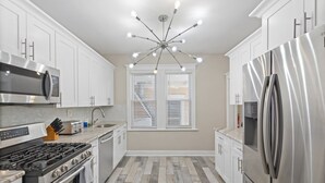 A modern kitchen with white cabinetry, stainless steel appliances, a central light fixture, and a window with blinds.