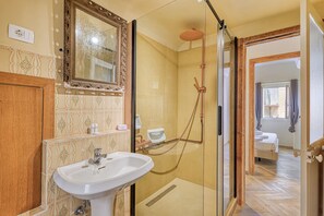 A bathroom with a sink, mirror, shower, and a bed visible through an open door. - Boutique Apartment Casa Del Giudice (Ventimiglia)