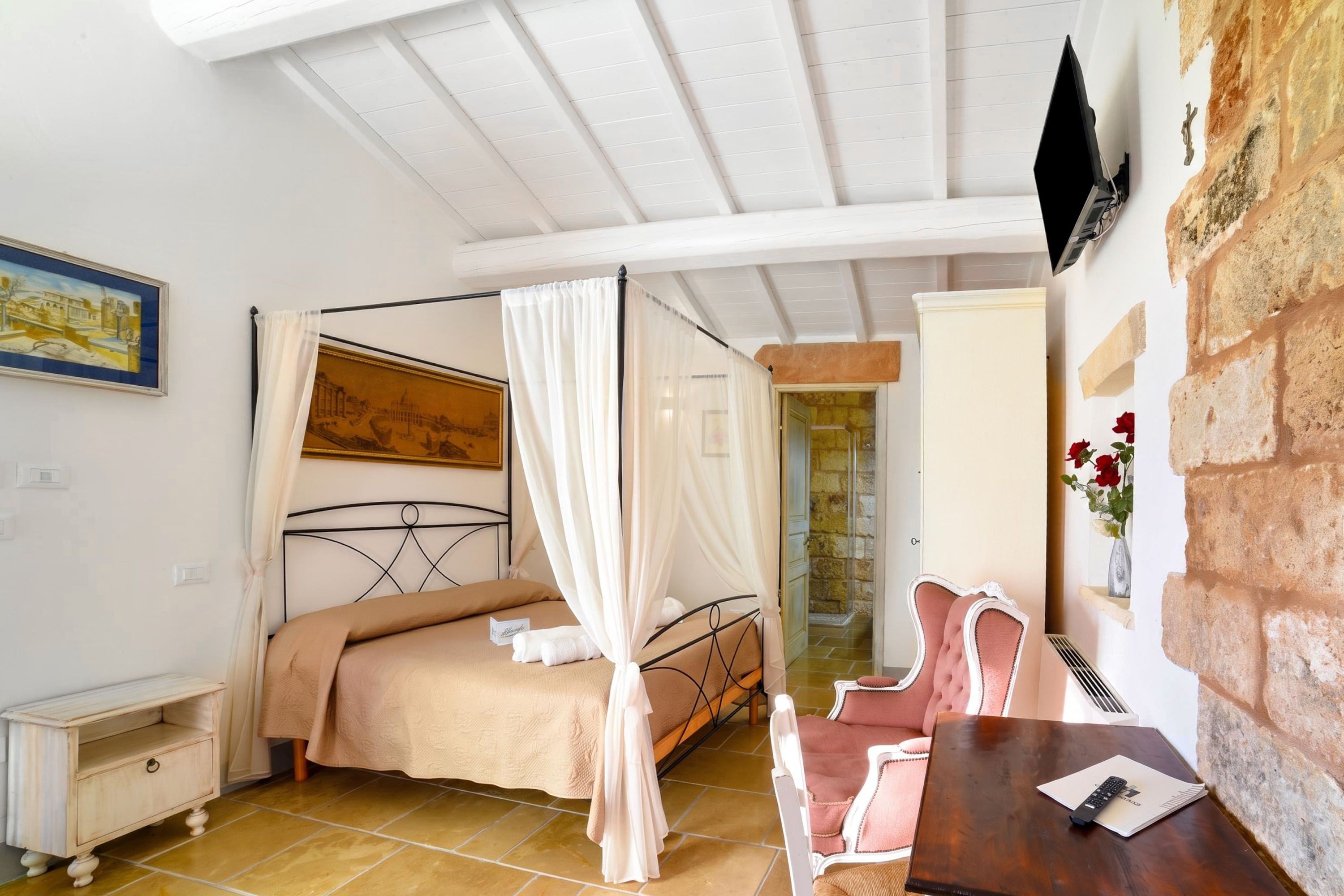 A bedroom with a four-poster bed, a bedside table, a chair, a desk, and a painting on the wall.