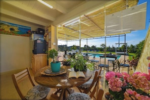 A covered patio with a pool, wooden furniture, and potted plants.