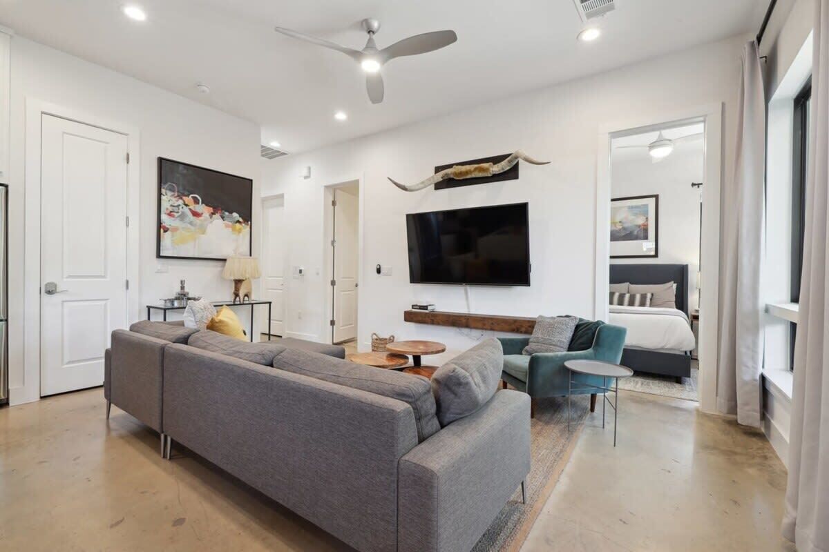 Un salon moderne avec un canapé gris, une petite table ronde, un téléviseur fixé au mur et un ventilateur de plafond.