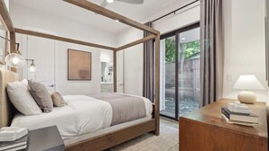A modern bedroom with a four-poster bed, a bedside table with books, a large window with curtains, and a view of greenery outside. - 4BR East Austin Home | Modern & Open Living (Austin)
