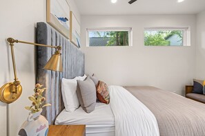 Un dormitorio con cama, mesita de noche, lámpara y ventanas.