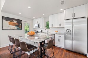 A modern kitchen with white cabinets, a stainless steel refrigerator, a marble island, and a painting on the wall. - Luxe 4BR Austin Home | Balcony & Private Deck (Austin)