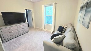 A room with a flat-screen TV on a dresser, a grey sofa with cushions, a window with curtains, and a door leading to another room.