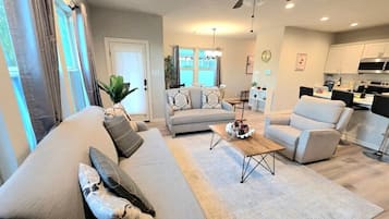 A modern living room with a sofa, armchairs, and a coffee table. There is a dining area with a table and chairs in the background.