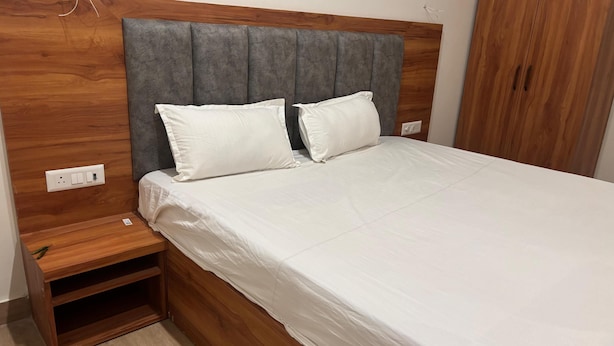 A neatly made bed with a wooden headboard and two pillows, a wooden nightstand with a power outlet, and a wardrobe with a wooden finish. - OnlyStay Surbhi Dham (Vrindavan)