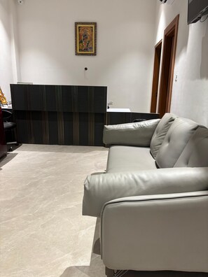 A modern living room with a beige leather sofa, a dark wooden cabinet, and a small white table. - OnlyStay Surbhi Dham (Vrindavan)
