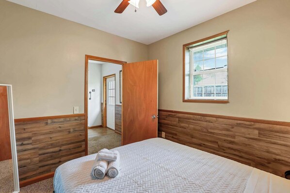 A bedroom with a wooden headboard, a bed with a quilt, a window with blinds, and a door leading to another room.