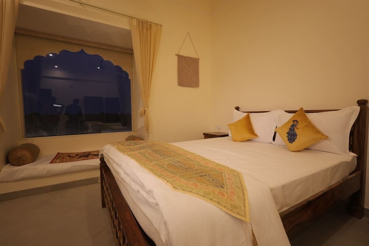 A hotel room with a large bed, two pillows, a bedside table, a nightstand, a window with curtains, and a wall-mounted hanging.