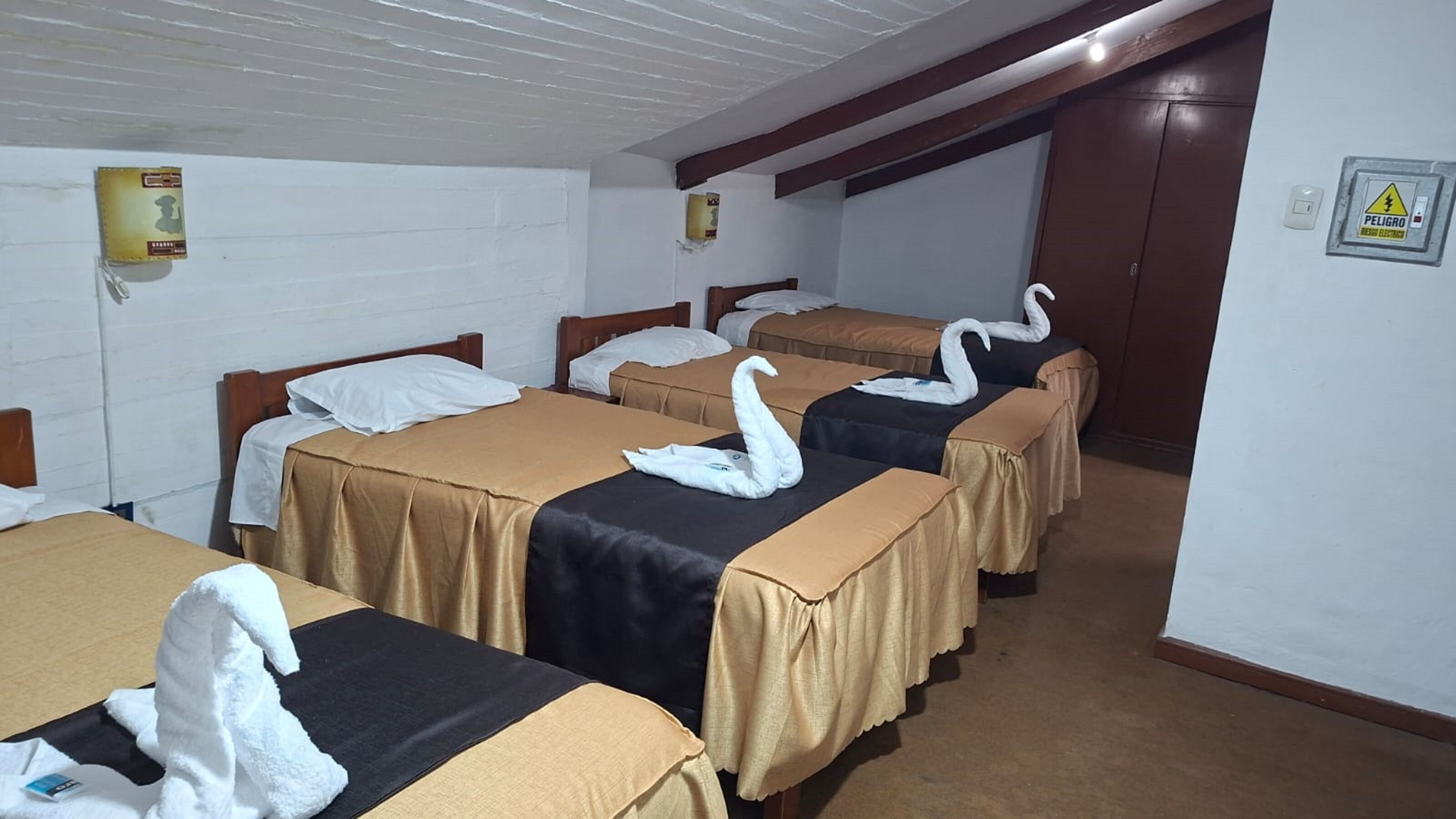 A hotel room with four beds, each with a swan-shaped towel. There are wall-mounted light switches and a warning sign on the wall.