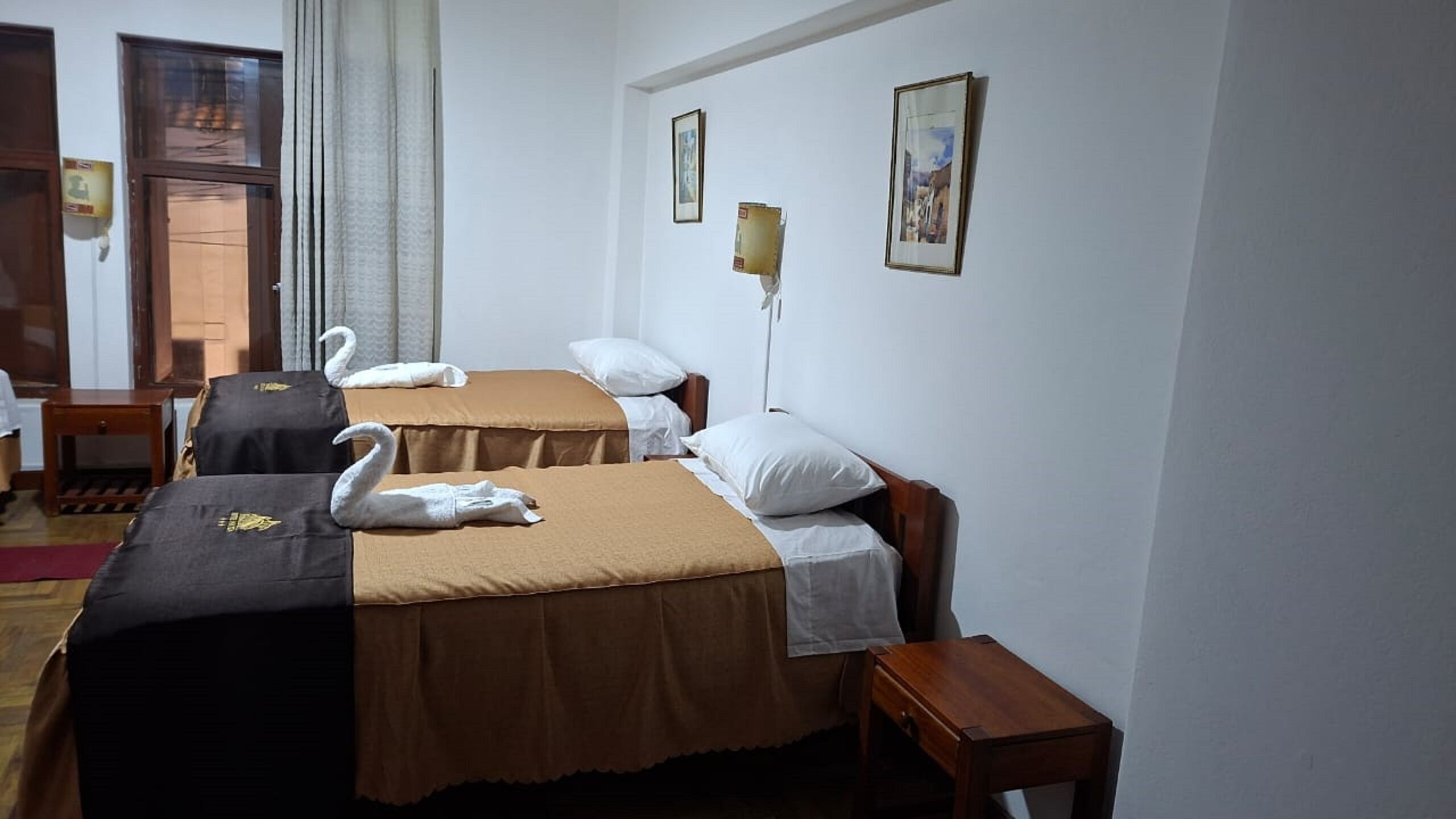 A hotel room with two beds, each with a brown bedspread and white pillows. There are two framed pictures on the wall, a wooden nightstand, and a window with curtains.