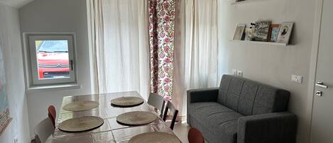 Comedor con una mesa preparada para cuatro, un sofá gris y una ventana con vista a un coche rojo.