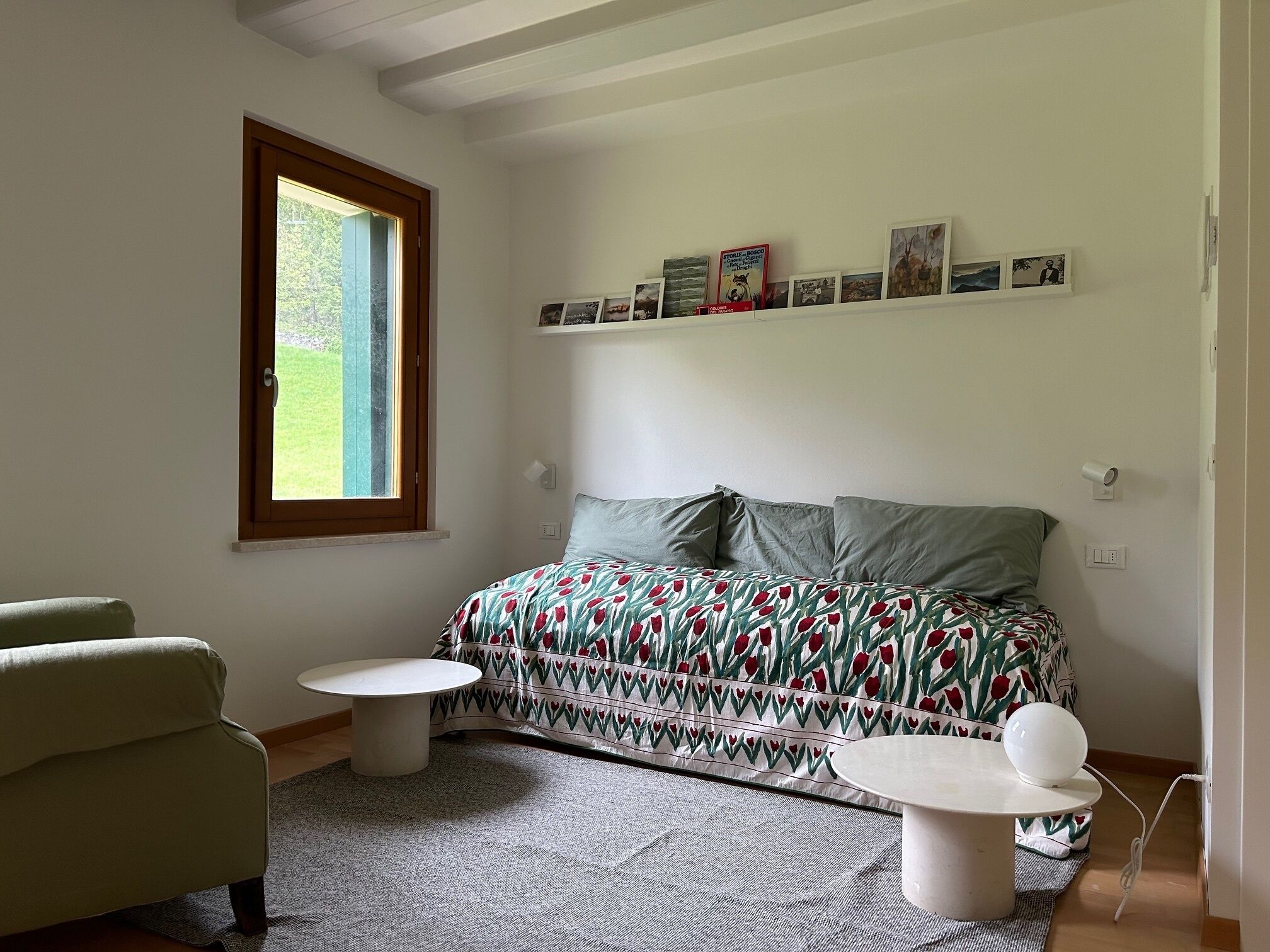 Ein Schlafzimmer mit einem Bett, einem kleinen runden Tisch, einem Stuhl, einem Fenster und einem Regal mit Büchern und Bildern.
