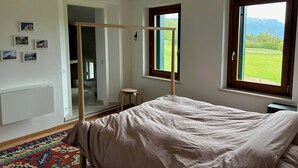 Une chambre avec un lit, une fenêtre donnant sur un champ verdoyant et des montagnes, un petit tabouret et un radiateur.