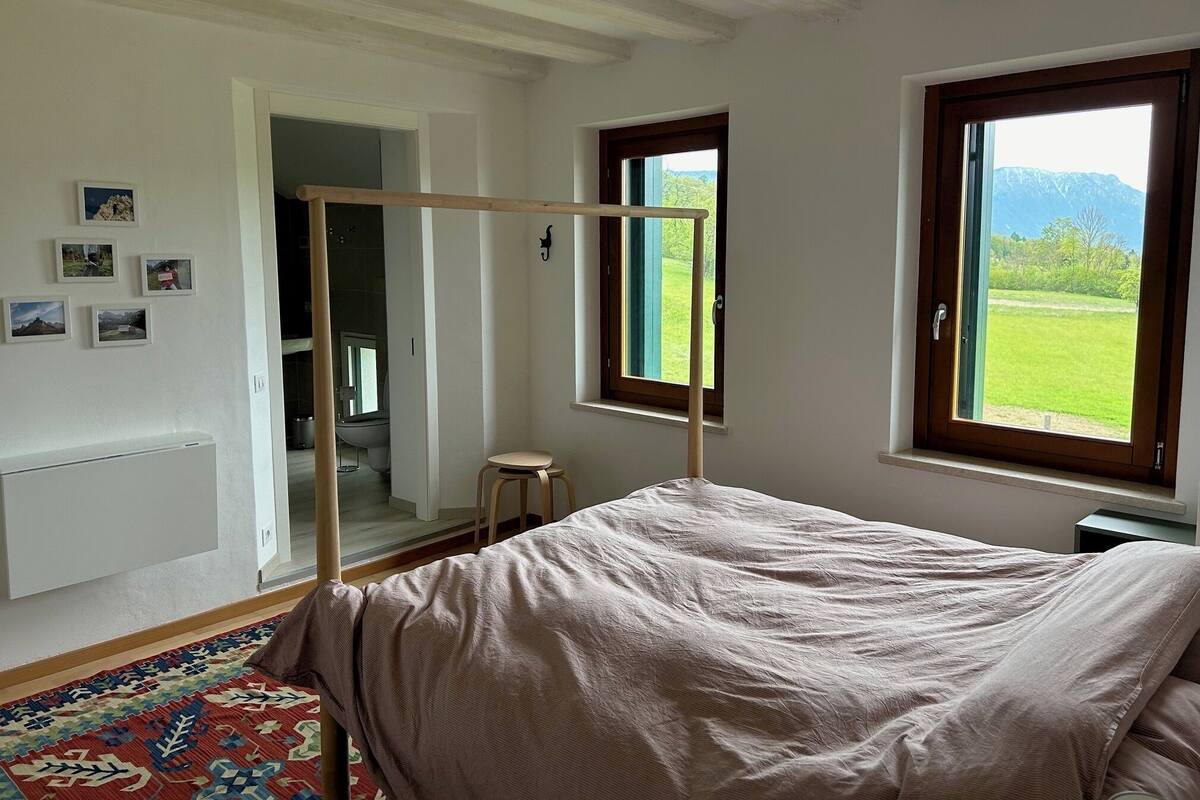 A bedroom with a bed, a window with a view of a green field and mountains, a small stool, and a radiator.