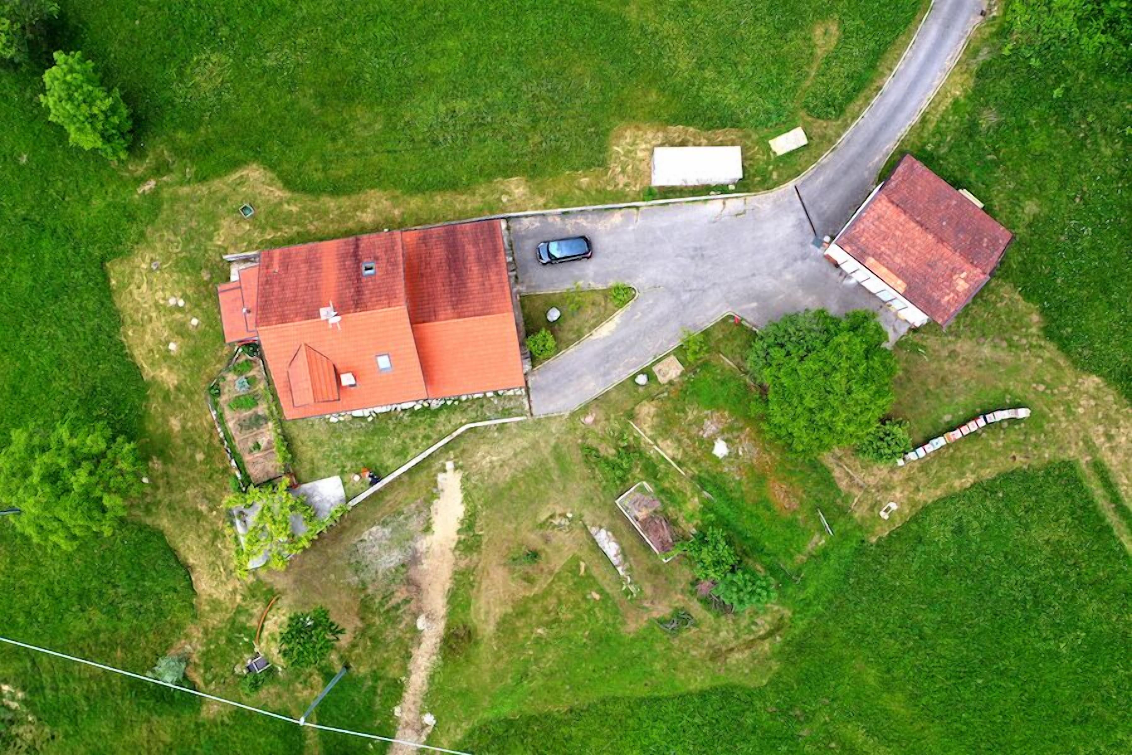 An aerial view of a property with a main building, a smaller attached structure, a paved driveway, and surrounding green fields.