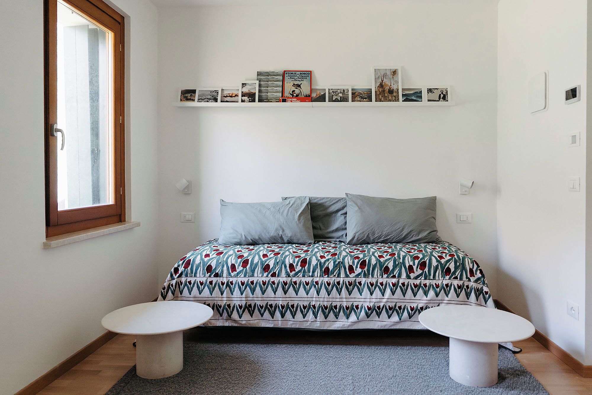 A bedroom with a bed, two bedside tables, a window, and a shelf with photos and books.