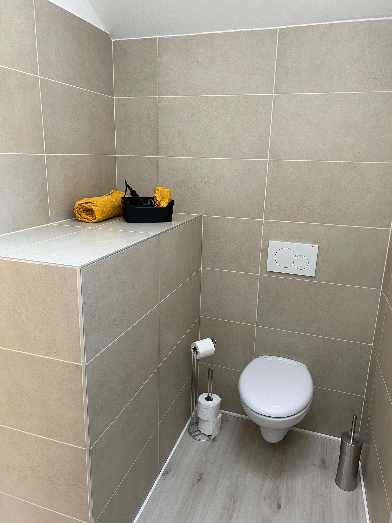 Een compacte, moderne badkamer met beige tegels, een wit toilet, een toiletrolhouder en een zwarte opbergkast met gele handdoeken.