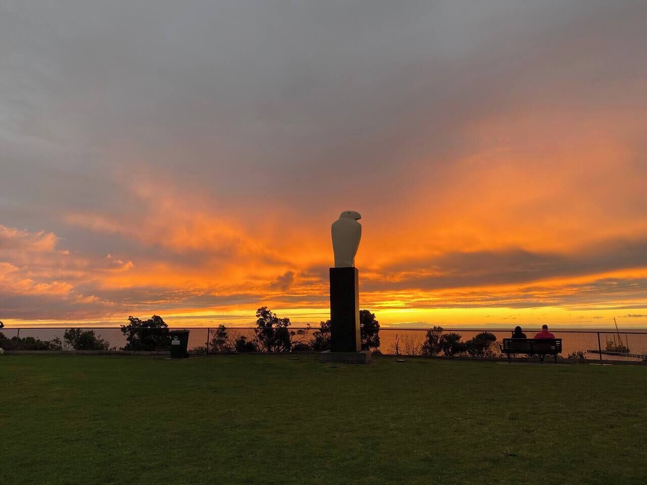A grassy area with a white sculpture on a pedestal, a fence, and a sunset in the background.