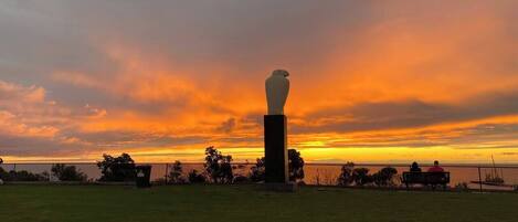A grassy area with a white sculpture on a pedestal, a fence, and a sunset in the background.