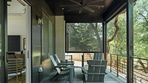 A covered outdoor seating area with metal chairs and a table, a television on the wall, and a view of trees through the glass.