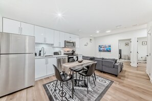 A modern kitchen with white cabinets, a stainless steel refrigerator, and a microwave. There is a dining area with a table and chairs, and a gray sofa in the living room. -  2-Bed, w/ In-Unit Washer & Dryer, Cozy Living,1-2 Min Drive to Anthony Henday! (Edmonton)