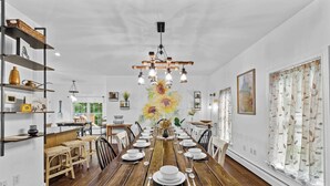 Un comedor con una larga mesa de madera dispuesta para una comida, rodeada de sillas. Hay estanterías con varios objetos a la izquierda, y un cuadro grande con motivos florales en la pared.
