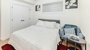 A bedroom with a bed, a chair, a small table, and a door. - Cozy 2-bedroom basement suite, fully furnished for comfort and convenience. (Edmonton)