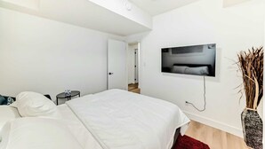A modern hotel room with a large bed, a small bedside table, a TV mounted on the wall, and a decorative vase. - Cozy 2-bedroom basement suite, fully furnished for comfort and convenience. (Edmonton)