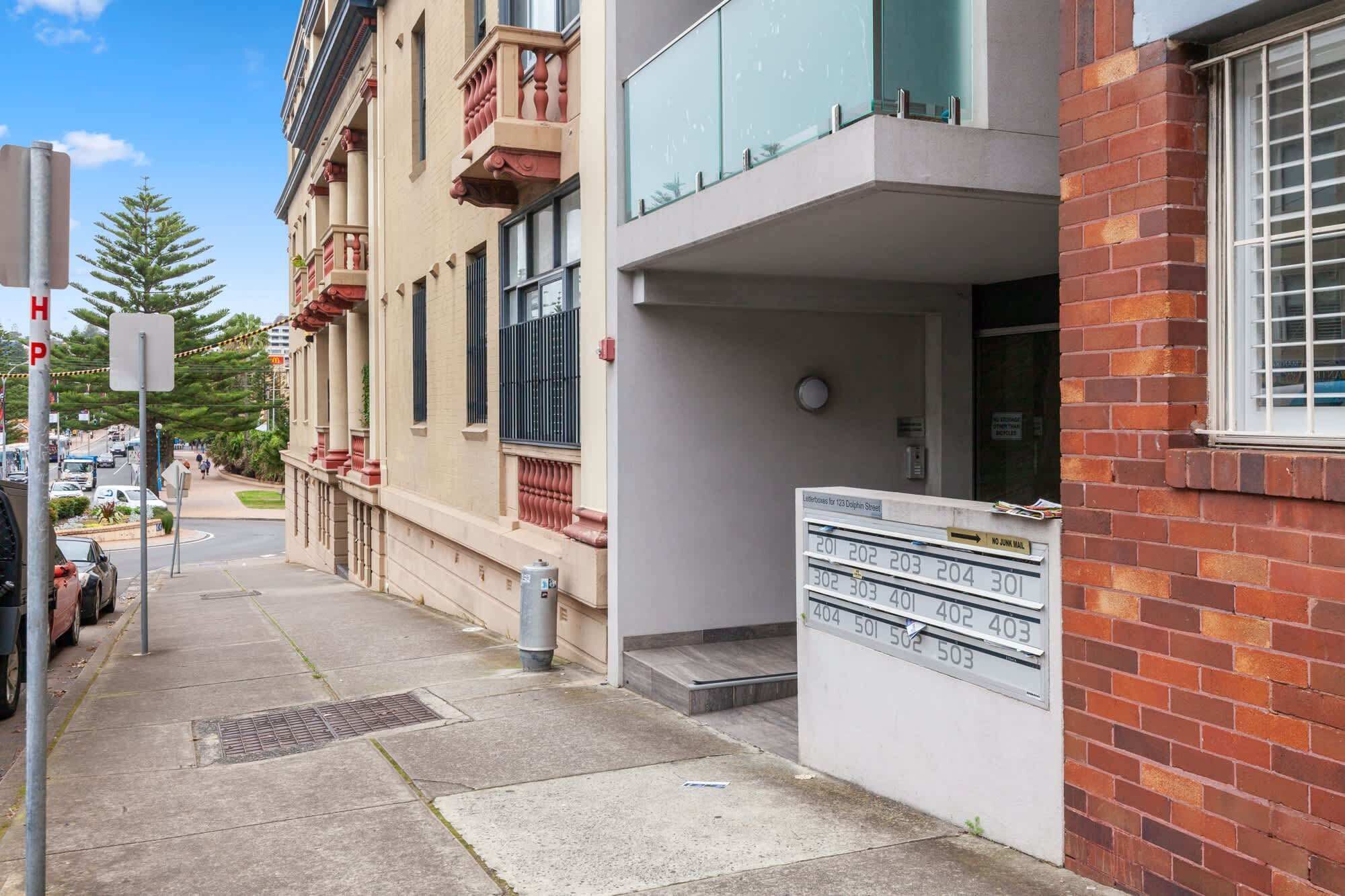 Une vue de rue montrant un immeuble doté d’un balcon avec des balustrades en verre, d’une boîte aux lettres et d’un panneau indiquant qu’il est interdit de déposer du courrier publicitaire.