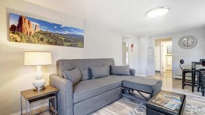 A modern living room with a grey sectional sofa, a small side table with a lamp, a coffee table with magazines, a large landscape painting, and a dining area with a table and chairs.