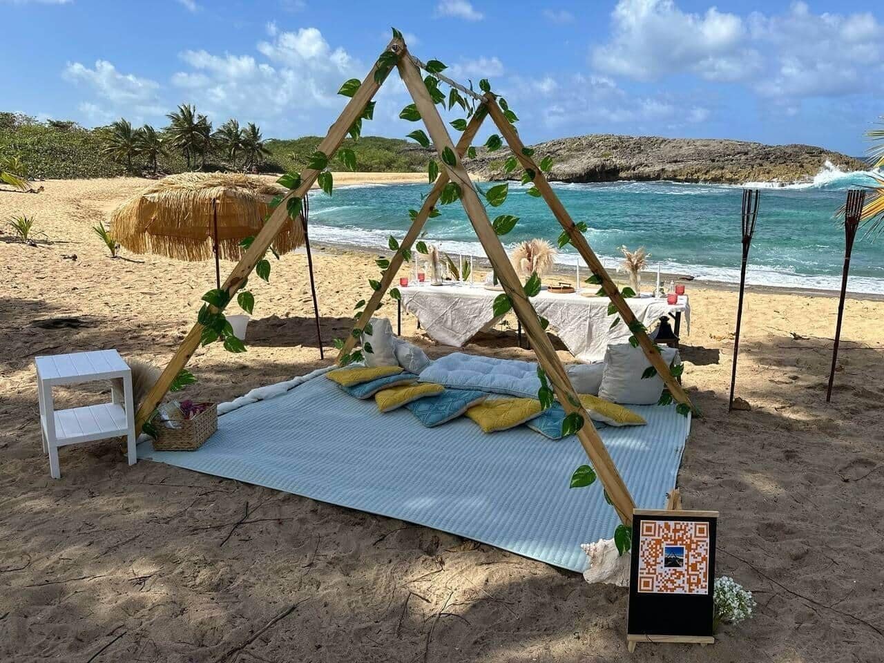 Een strandopstelling met een houten A-frame constructie, een witte bank, een picknicktafel en een standaard voor een QR-code.