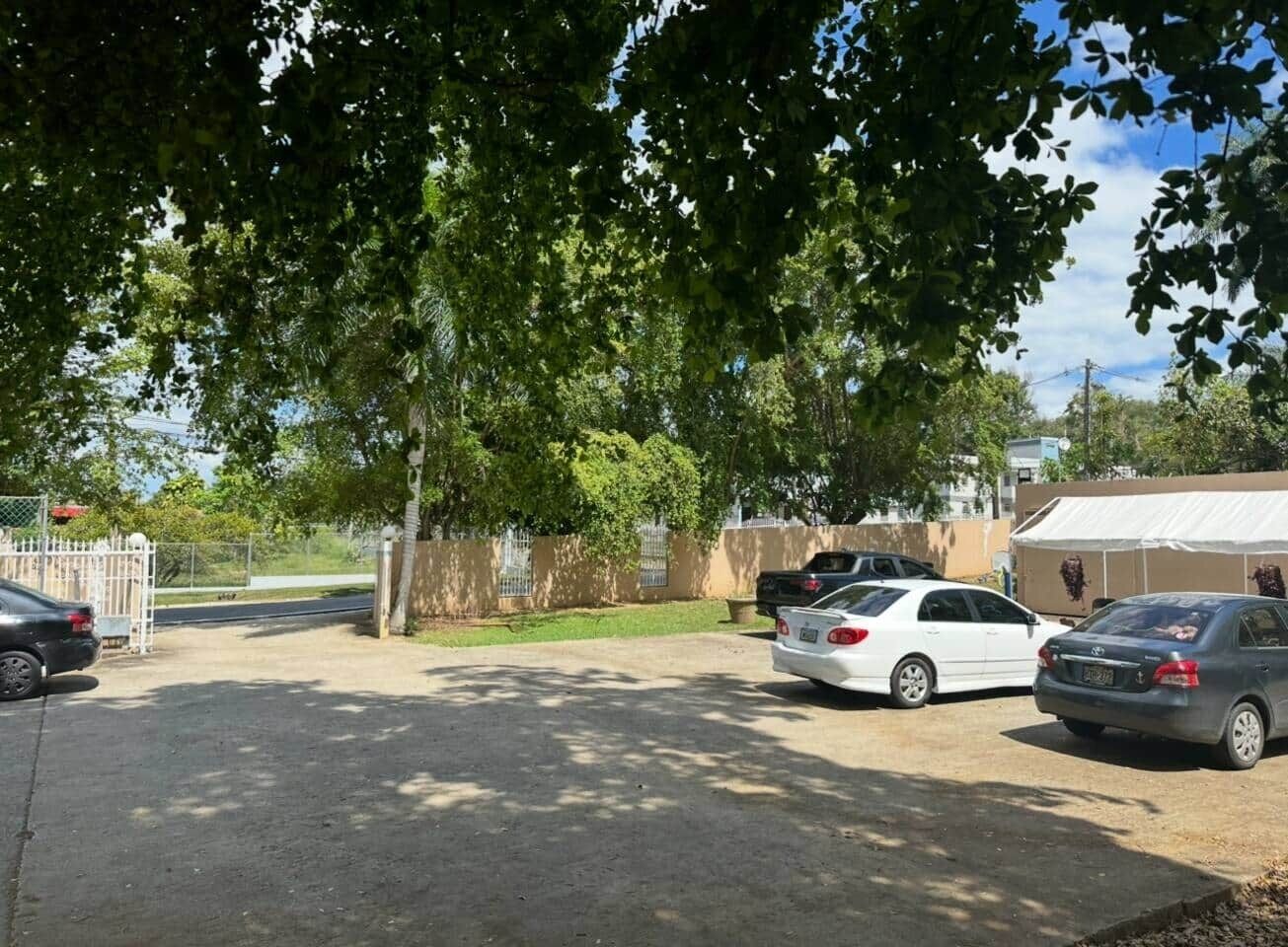 Un estacionamiento con coches, una entrada con portón y un edificio con techo blanco al fondo.